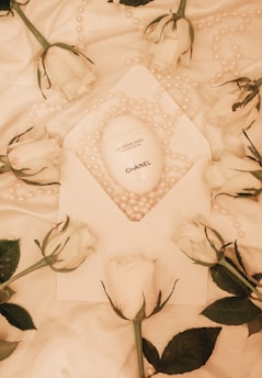 An arrangement features a white Chanel hand cream product placed on a white fabric amidst scattered pearl beads and several white roses. The elegant composition suggests a luxurious and refined aesthetic.