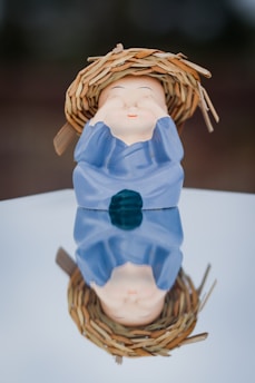 A ceramic figurine of a cheerful character wearing a blue robe and a straw hat is placed on a reflective surface. Its facial features suggest a playful expression as it covers its eyes with its hands. The reflection is clearly visible on the shiny surface below.