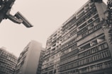 A black and white photograph featuring tall residential buildings with numerous balconies, creating an urban landscape. A large security camera is prominently positioned in the top left corner, overlooking the scene. The buildings have a vintage and weathered appearance, with visible variations in the windows and balcony structures. Sparse foliage is seen at the edges, adding a hint of nature.