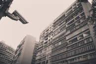 A black and white photograph featuring tall residential buildings with numerous balconies, creating an urban landscape. A large security camera is prominently positioned in the top left corner, overlooking the scene. The buildings have a vintage and weathered appearance, with visible variations in the windows and balcony structures. Sparse foliage is seen at the edges, adding a hint of nature.