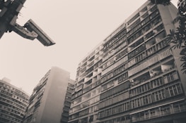 A black and white photograph featuring tall residential buildings with numerous balconies, creating an urban landscape. A large security camera is prominently positioned in the top left corner, overlooking the scene. The buildings have a vintage and weathered appearance, with visible variations in the windows and balcony structures. Sparse foliage is seen at the edges, adding a hint of nature.