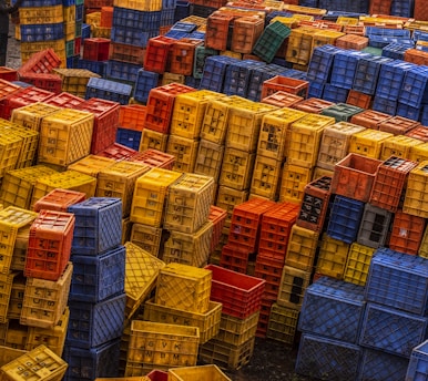 A large number of colorful plastic crates are stacked in an outdoor setting, creating a patchwork of colors and patterns. The crates are mostly yellow, red, blue, and orange, arranged in an almost chaotic yet organized manner. Some crates have letters and symbols marked on them.