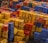 A large number of colorful plastic crates are stacked in an outdoor setting, creating a patchwork of colors and patterns. The crates are mostly yellow, red, blue, and orange, arranged in an almost chaotic yet organized manner. Some crates have letters and symbols marked on them.