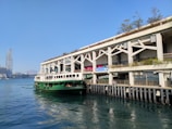 A green ferry is docked at a pier, adjacent to a modern building with a green roof and plants. In the background, a tall skyscraper rises against a clear blue sky. The water is calm and reflects the surroundings.