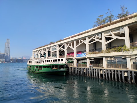A green ferry is docked at a pier, adjacent to a modern building with a green roof and plants. In the background, a tall skyscraper rises against a clear blue sky. The water is calm and reflects the surroundings.