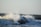 Waves crashing against large rocks, creating a spray of water droplets. The ocean appears turbulent with a series of waves continuing into the horizon under a clear sky.