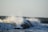 Waves crashing against large rocks, creating a spray of water droplets. The ocean appears turbulent with a series of waves continuing into the horizon under a clear sky.