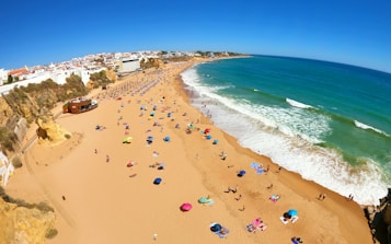 A wide sandy beach filled with colorful umbrellas and beachgoers enjoying the sun. The beach curves alongside the turquoise ocean, with waves gently lapping upon the shore. In the distance, a town with white buildings is visible atop golden cliffs.