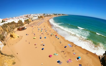 A wide sandy beach filled with colorful umbrellas and beachgoers enjoying the sun. The beach curves alongside the turquoise ocean, with waves gently lapping upon the shore. In the distance, a town with white buildings is visible atop golden cliffs.