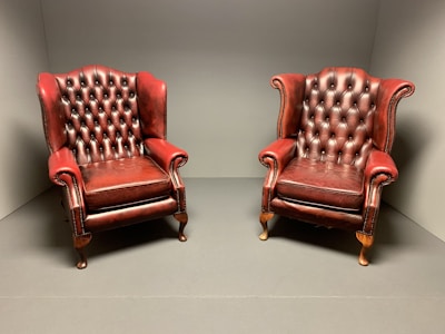 Two vintage, red leather armchairs with tufted backs sit side by side in a minimalist, neutral-colored room. The leather is slightly worn, adding a classic, lived-in feel.