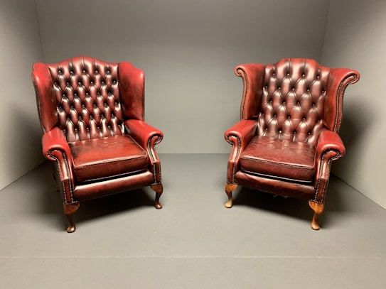 Two vintage, red leather armchairs with tufted backs sit side by side in a minimalist, neutral-colored room. The leather is slightly worn, adding a classic, lived-in feel.