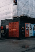 A construction site with scaffolding covered in white sheeting. The corner of the site features a black wall with repeated red text 'PAST PRESENT' and a singular 'FUTURE'. Various informational and advertisement boards are visible, promoting Clerys Quarter with images and text about work and catching up.