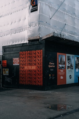 A construction site with scaffolding covered in white sheeting. The corner of the site features a black wall with repeated red text 'PAST PRESENT' and a singular 'FUTURE'. Various informational and advertisement boards are visible, promoting Clerys Quarter with images and text about work and catching up.