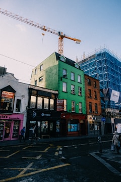 A city street with colorful buildings, including a prominent green building with shops at street level. A large construction crane is visible towering above, with a nearby building covered in scaffolding. Pedestrians walk along the sidewalk, and various shop signs are displayed.