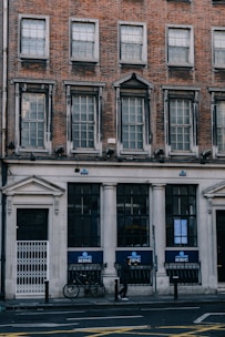 A brick building with large windows and a classic architectural style. The ground level features a bank with the KBC logo visible on the signage. A bicycle is parked against the building, and a person is walking on the sidewalk. The structure has a historic aesthetic with multiple rows of windows.