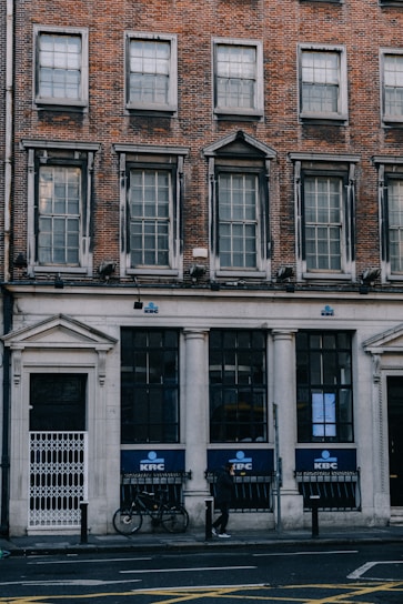 A brick building with large windows and a classic architectural style. The ground level features a bank with the KBC logo visible on the signage. A bicycle is parked against the building, and a person is walking on the sidewalk. The structure has a historic aesthetic with multiple rows of windows.