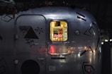 A close-up view of a metal trailer with geometric and floral decals. Through a window, soft yellow light illuminates a refrigerator stocked with beverages, notably Coca-Cola cans and bottles.