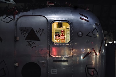 A close-up view of a metal trailer with geometric and floral decals. Through a window, soft yellow light illuminates a refrigerator stocked with beverages, notably Coca-Cola cans and bottles.