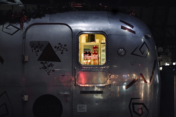 A close-up view of a metal trailer with geometric and floral decals. Through a window, soft yellow light illuminates a refrigerator stocked with beverages, notably Coca-Cola cans and bottles.