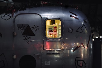 A close-up view of a metal trailer with geometric and floral decals. Through a window, soft yellow light illuminates a refrigerator stocked with beverages, notably Coca-Cola cans and bottles.
