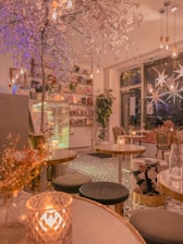 A cozy cafe interior with tables and chairs adorned with floral patterns. Warm lighting from pendant lights and candles illuminates the space. A decorative tree with pink blossoms hangs from the ceiling. Shelves with books and potted plants are visible along one wall. The floor has intricate black and white tile patterns.