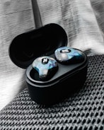 A pair of wireless earbuds resting in an open charging case. The earbuds have a unique, colorful design with a textured pattern and a small logo. They are placed on a textured black surface, with a soft, light-colored drape in the background.