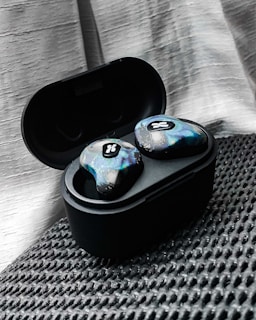 A pair of wireless earbuds resting in an open charging case. The earbuds have a unique, colorful design with a textured pattern and a small logo. They are placed on a textured black surface, with a soft, light-colored drape in the background.