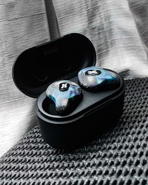 A pair of wireless earbuds resting in an open charging case. The earbuds have a unique, colorful design with a textured pattern and a small logo. They are placed on a textured black surface, with a soft, light-colored drape in the background.