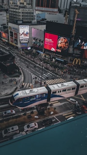 An urban scene featuring a busy city intersection with a modern light rail train passing overhead. The streets are bustling with traffic, including cars and pedestrians crossing the road. Large digital billboards display advertisements on the buildings surrounding the intersection. Notable establishments such as McDonald's and Starbucks are visible among the buildings. The architectural style is contemporary with high-rise buildings in the background.