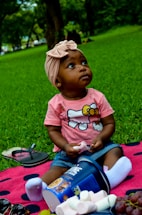 A toddler wearing a pink shirt with a cartoon character sits on a pink and black polka-dotted blanket in a grassy area. The child wears a light pink headband and white socks, holding a marshmallow. An overturned container of cereal is next to the child, with spilled marshmallows and a bunch of grapes nearby. Flip-flops and sunglasses lie on the blanket. Trees and green foliage are visible in the background.
