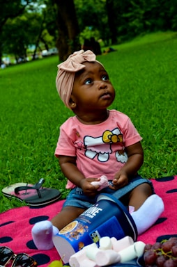 A toddler wearing a pink shirt with a cartoon character sits on a pink and black polka-dotted blanket in a grassy area. The child wears a light pink headband and white socks, holding a marshmallow. An overturned container of cereal is next to the child, with spilled marshmallows and a bunch of grapes nearby. Flip-flops and sunglasses lie on the blanket. Trees and green foliage are visible in the background.