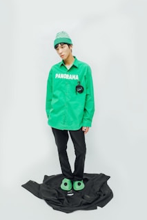 A person stands against a minimalist background wearing a bright green beanie and a green shirt with the word 'PANORAMA' printed on it. The person is also wearing black pants and green slippers. A black garment or fabric is placed on the floor underneath their feet.