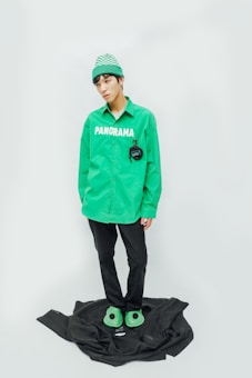 A person stands against a minimalist background wearing a bright green beanie and a green shirt with the word 'PANORAMA' printed on it. The person is also wearing black pants and green slippers. A black garment or fabric is placed on the floor underneath their feet.