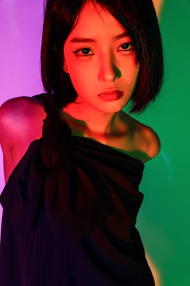 A woman with short black hair stands against a vivid multicolored background, creating a dramatic lighting effect. Her expression is serious, and she is wearing an off-the-shoulder black outfit. The lighting casts shades of red, green, and pink across her face and body.