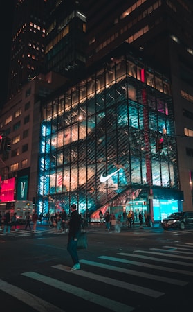 A bustling urban scene at night featuring a large, well-lit storefront with a prominent Nike logo. The building's glass facade is illuminated with vibrant teal and red lights, reflecting the hustle and bustle of people on the street below. A crosswalk is visible in the foreground with pedestrians crossing, alongside a parked bus and moving cars. The towering buildings create a sense of height and energy in the cityscape.