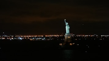 A night scene featuring the illuminated Statue of Liberty in the foreground, set against a backdrop of numerous city lights twinkling across the horizon. The dark sky contrasts with the glowing statue and lights, creating a striking and iconic view.