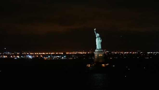 A night scene featuring the illuminated Statue of Liberty in the foreground, set against a backdrop of numerous city lights twinkling across the horizon. The dark sky contrasts with the glowing statue and lights, creating a striking and iconic view.