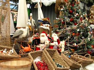A festive scene featuring a decorated Christmas tree with ornaments and tinsel. Several Santa Claus figurines are positioned near the tree, surrounded by wicker baskets. There are also nautical elements like model sailboats and pelicans. A lifelike crab model adds a playful touch to the display.