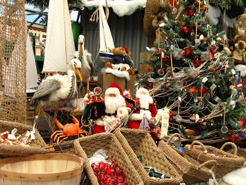 A festive scene featuring a decorated Christmas tree with ornaments and tinsel. Several Santa Claus figurines are positioned near the tree, surrounded by wicker baskets. There are also nautical elements like model sailboats and pelicans. A lifelike crab model adds a playful touch to the display.