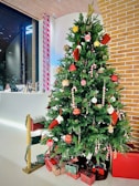 A decorated Christmas tree adorned with ornaments including red and gold baubles, candy canes, and small stockings, standing beside a white wall and exposed brick structure. Underneath the tree are several wrapped gifts in red and green packaging, and a small mailbox attached to a post decorated with garland.
