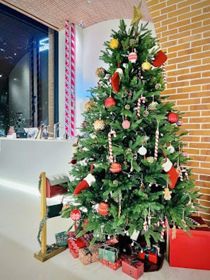 A decorated Christmas tree adorned with ornaments including red and gold baubles, candy canes, and small stockings, standing beside a white wall and exposed brick structure. Underneath the tree are several wrapped gifts in red and green packaging, and a small mailbox attached to a post decorated with garland.