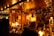 A festive market stall adorned with warm string lights and various decorative ornaments. The stall is filled with handmade crafts, holiday decorations, and seasonal items. There is a person in a winter coat browsing the offerings.