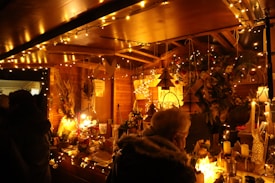 A festive market stall adorned with warm string lights and various decorative ornaments. The stall is filled with handmade crafts, holiday decorations, and seasonal items. There is a person in a winter coat browsing the offerings.