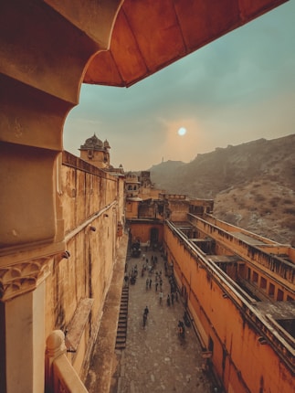 A historic fort with old architecture featuring ornate details and warm colors. The setting sun casts an orange hue over the scene, highlighting the intricate structures and the long narrow pathway below with a group of people walking. The backdrop includes hilly terrain under a cloudy sky.
