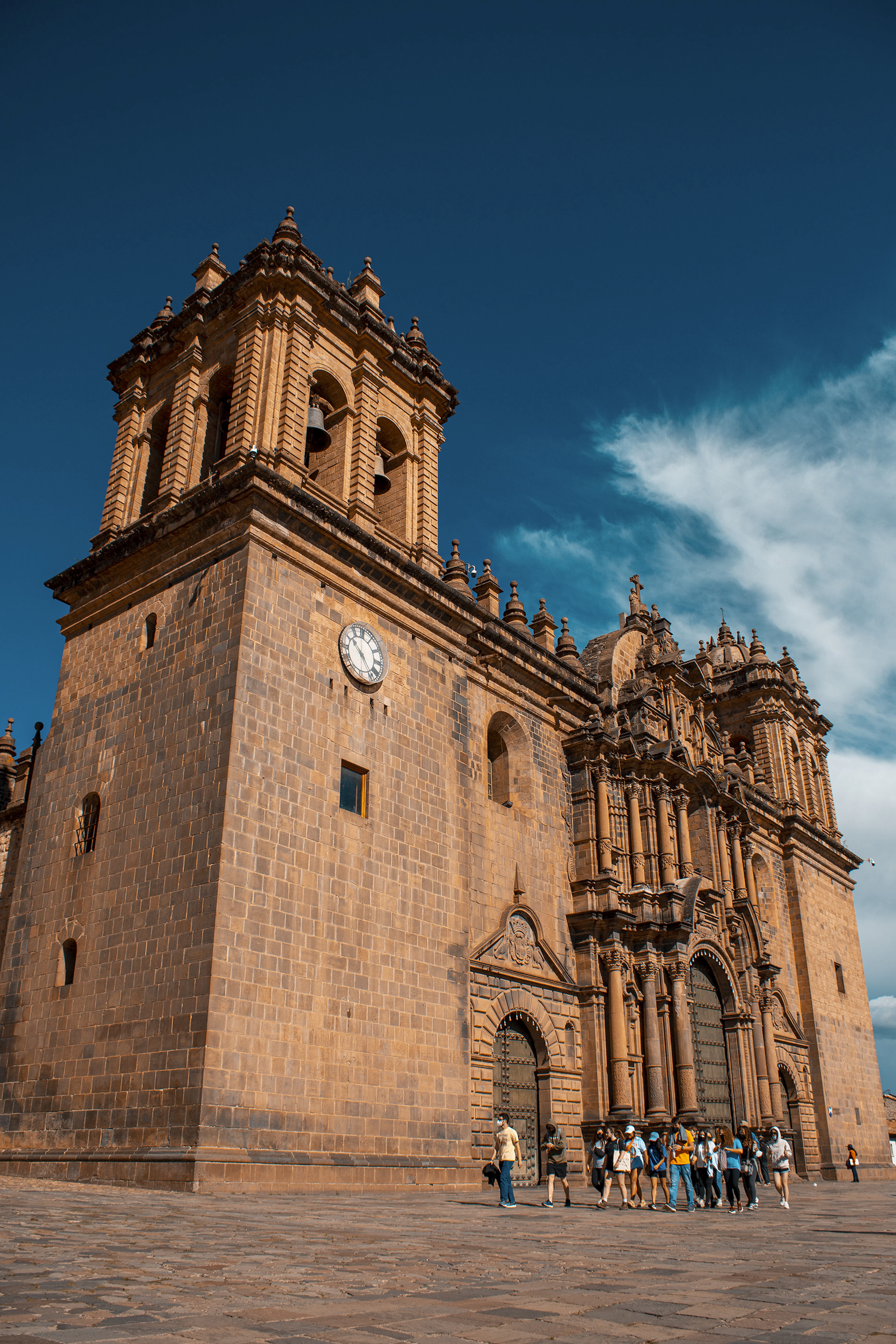 Cusco's cathedral bell tower rises against a deep blue sky, tourists gathered at its base.