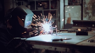 A person wearing a welding helmet is engaged in a welding task, with bright sparks flying from the point of contact on a metal surface. The setting appears to be a workshop with various tools and materials on a table.