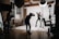 A well-lit photography studio with a model posing in a black, form-fitting outfit adorned with reflective elements. The photographer is capturing the scene with professional equipment, including lights and backdrops. The studio has a large window, wooden floors, and brick walls, creating a warm and creative atmosphere.
