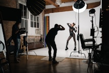 A well-lit photography studio with a model posing in a black, form-fitting outfit adorned with reflective elements. The photographer is capturing the scene with professional equipment, including lights and backdrops. The studio has a large window, wooden floors, and brick walls, creating a warm and creative atmosphere.