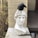 A white marble bust of a classical figure with a detailed, serene expression is positioned against a beige brick wall. Atop the bust's head, a black bird is perched. The setting includes some dry foliage and a wooden deck in the background. A small lantern-style decoration is placed near the base of the bust.
