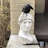 A white marble bust of a classical figure with a detailed, serene expression is positioned against a beige brick wall. Atop the bust's head, a black bird is perched. The setting includes some dry foliage and a wooden deck in the background. A small lantern-style decoration is placed near the base of the bust.